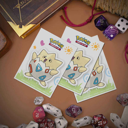 Tre Togepi-sleeves lagt ut på et spillbord sammen med terninger