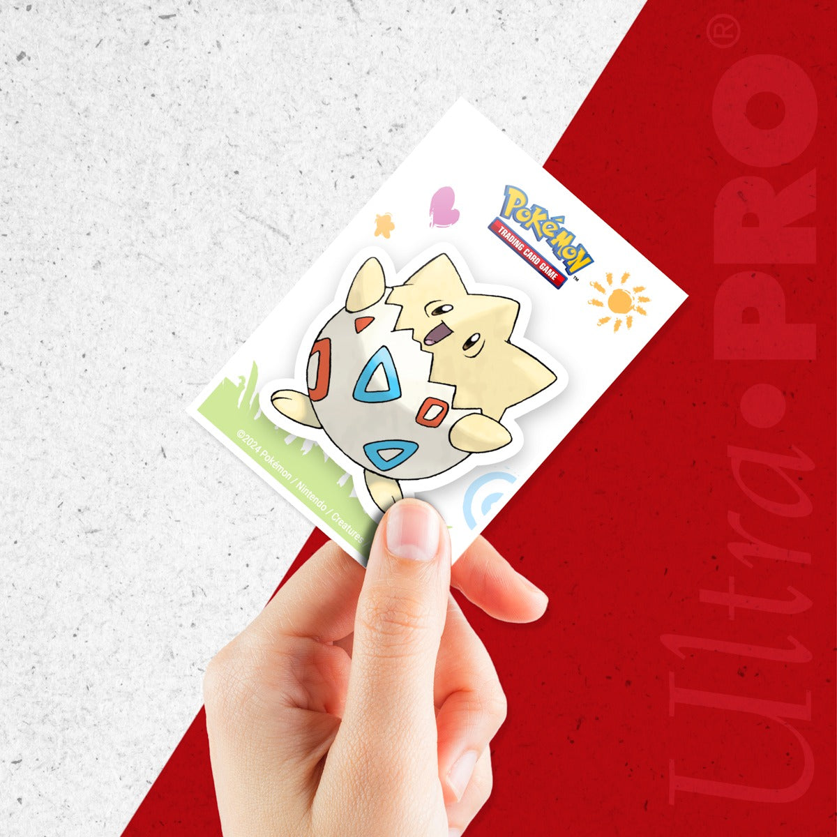 Hånd som holder en Togepi-sleeve fra Ultra Pro, med sol, hjerte og fargerike tegninger rundt Togepi
